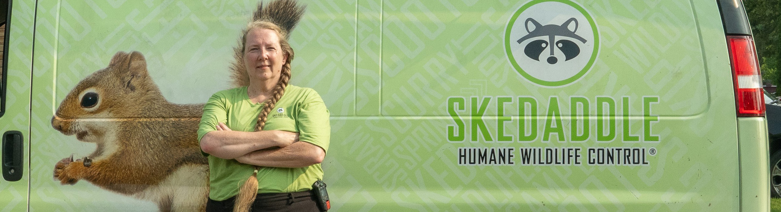 A female Skedaddle wildlife technician standing proudly in front of a green company service van with a squirrel illustration.
