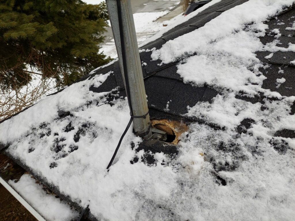 A large hole chewed by squirrels in roof shingles and plywood decking next to an electrical service mast covered in snow.