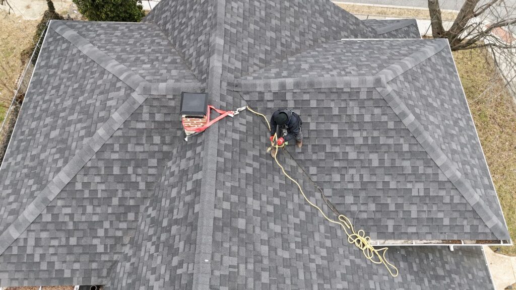 Aerial View of Roof Safety and Wildlife Exclusion Prep Overhead drone shot of a technician in a harness anchored to a chimney while working on a grey shingled roof.