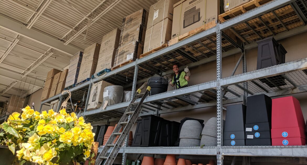 A technician in a safety harness giving a thumbs up from a high shelf in a large commercial warehouse.