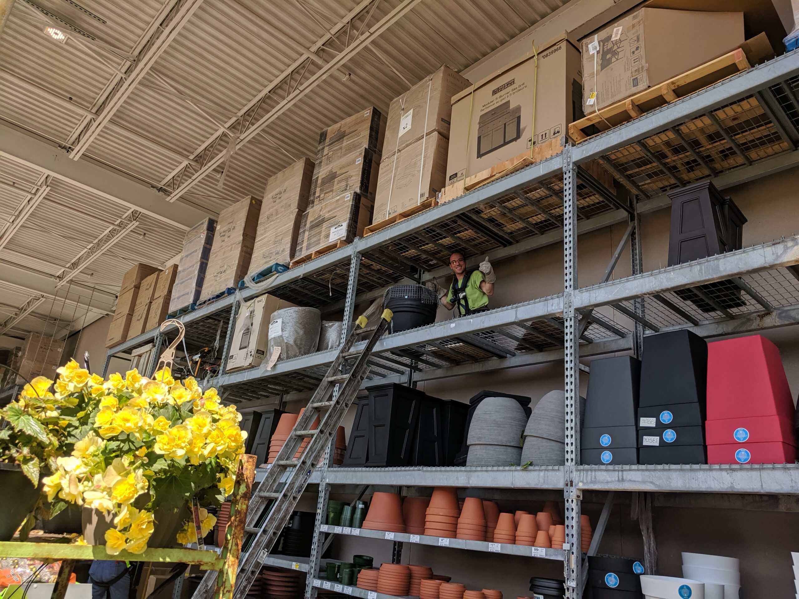 A technician in a safety harness giving a thumbs up from a high shelf in a large commercial warehouse.