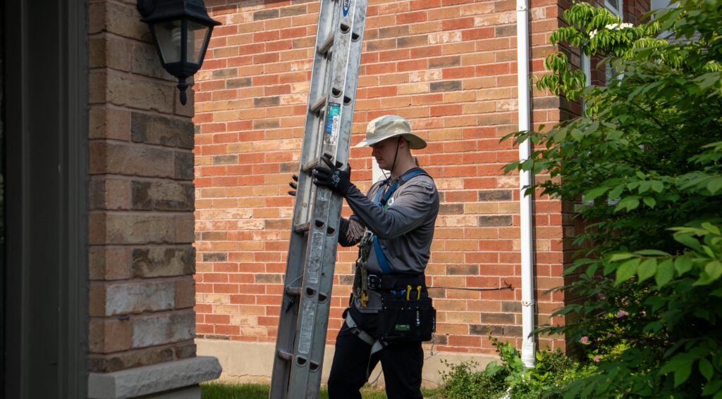 A Skedaddle technician wearing a safety harness, tool belt, and sun hat while securing a tall extension ladder against a brick house.