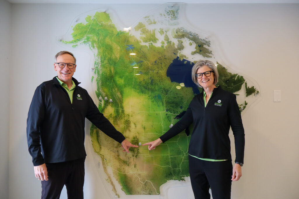 A Skedaddle technician uses a flashlight to inspect a dense bird's nest composed of dried grass and twigs inside a white exterior exhaust vent on a brick wall.