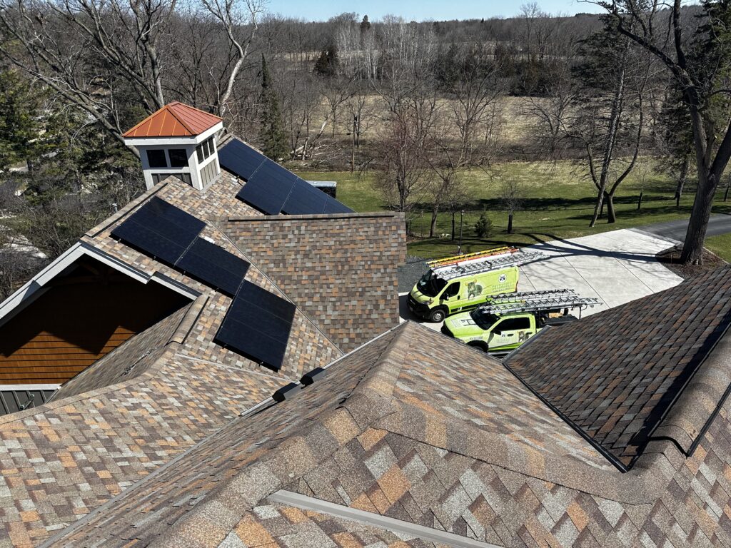 High-angle view of a large residential roof with solar panels and two Skedaddle service vehicles parked in the driveway below.
