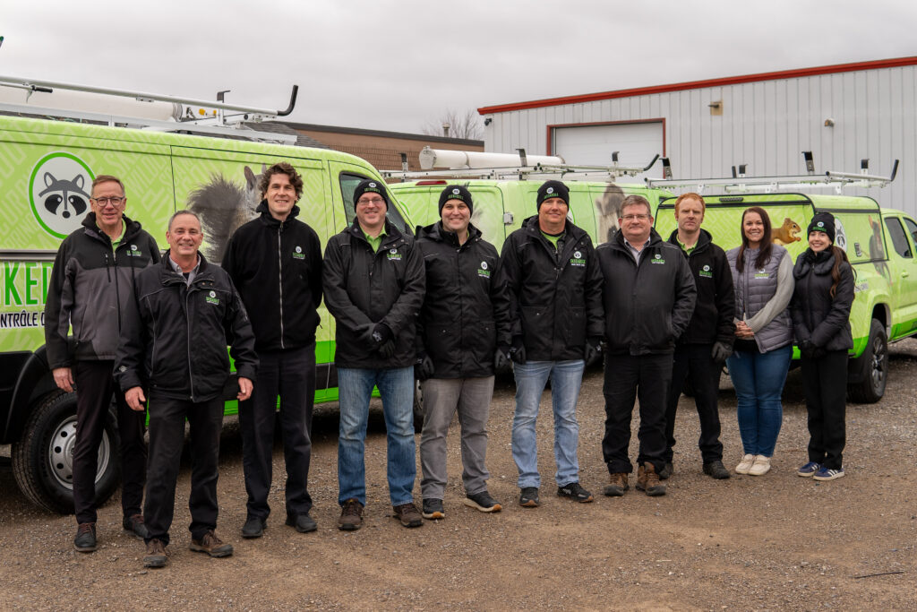 A group of ten Skedaddle Humane Wildlife Control team members in professional black and grey winter gear standing in front of three branded green service vehicles in Minneapolis, Minnesota.