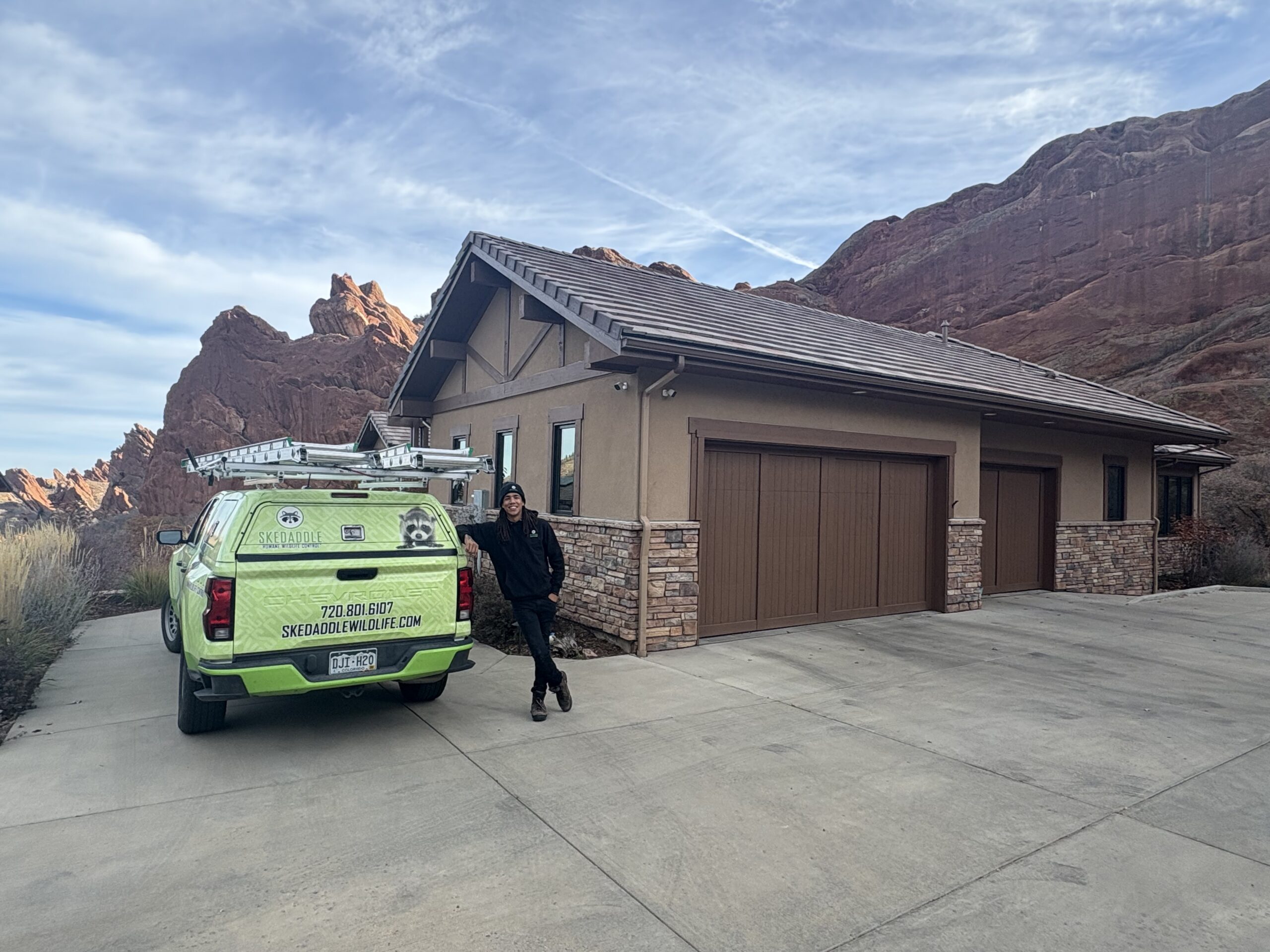 A smiling Skedaddle wildlife technician leaning against a lime green branded service truck parked in the driveway of a modern mountain home in Denver, Colorado.