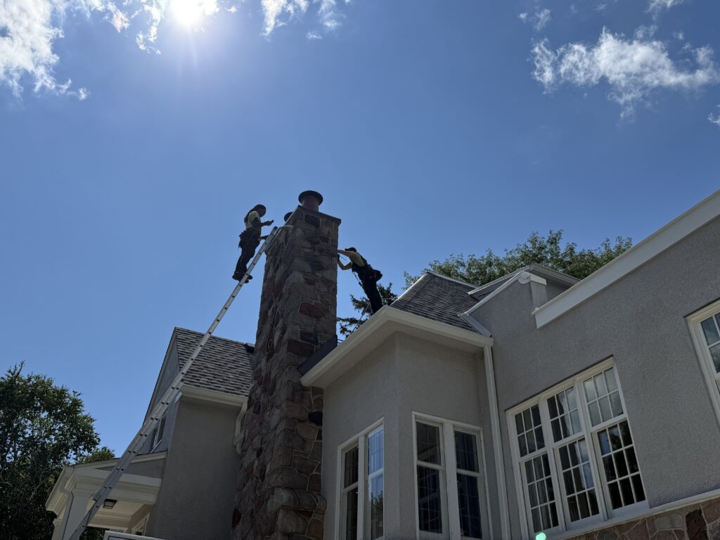 Skedaddle Technicians Performing Chimney Exclusion in Minneapolis Two Skedaddle Humane Wildlife Control technicians on ladders working at the top of a tall stone chimney on a sunny day.