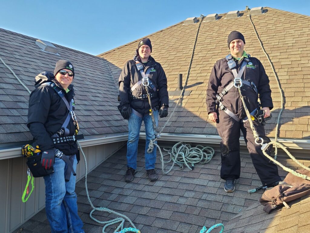 Skedaddle Wildlife Technicians Using Roof Safety Harnesses in Hennepin County Three Skedaddle Humane Wildlife Control technicians wearing winter gear and safety harnesses standing on a steep residential roof secured by ropes.