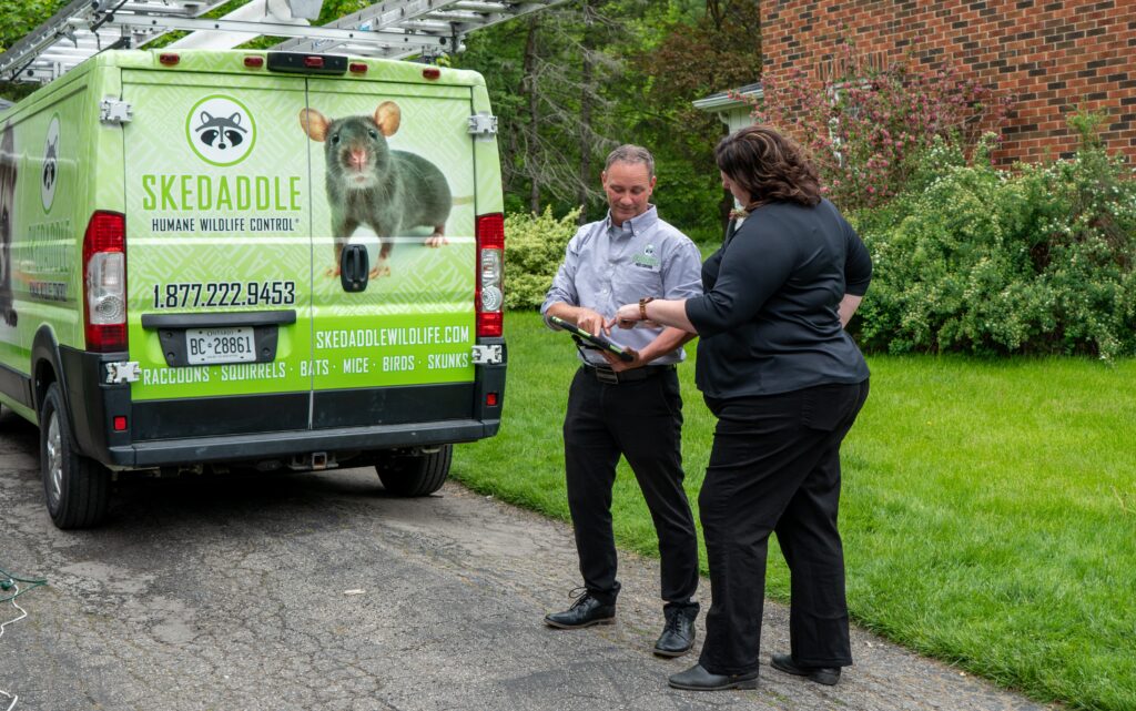 A Skedaddle technician and a homeowner standing behind a branded service van while reviewing inspection findings on a tablet.