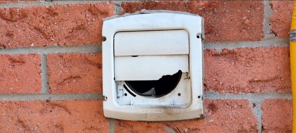 A white plastic louvered dryer vent cover on a red brick wall with a large hole chewed through the bottom flap, allowing wildlife access to the vent pipe.