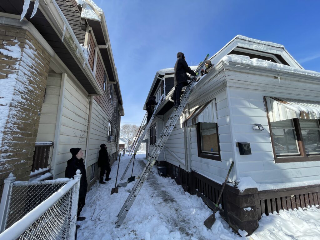 Skedaddle technicians using long ladders to inspect the roofline and gutters of a snow-covered white house.