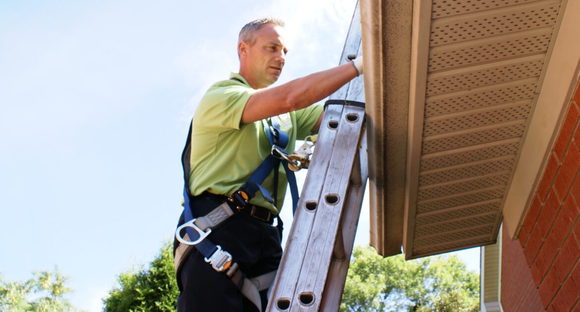 A Skedaddle technician wearing a safety harness climbs a ladder to inspect a home's soffit and gutters for squirrel entry points.