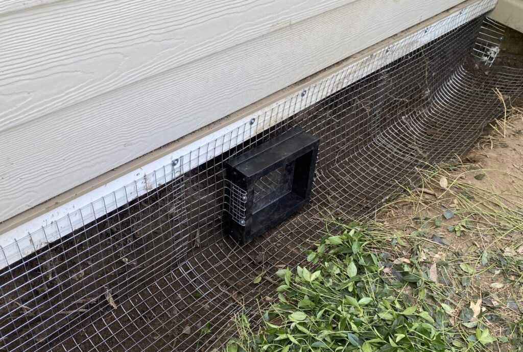 Humane wildlife exclusion mesh installation Galvanized steel hardware cloth mesh installed along the base of a house siding to prevent wildlife entry, featuring a specialized one-way door exit.