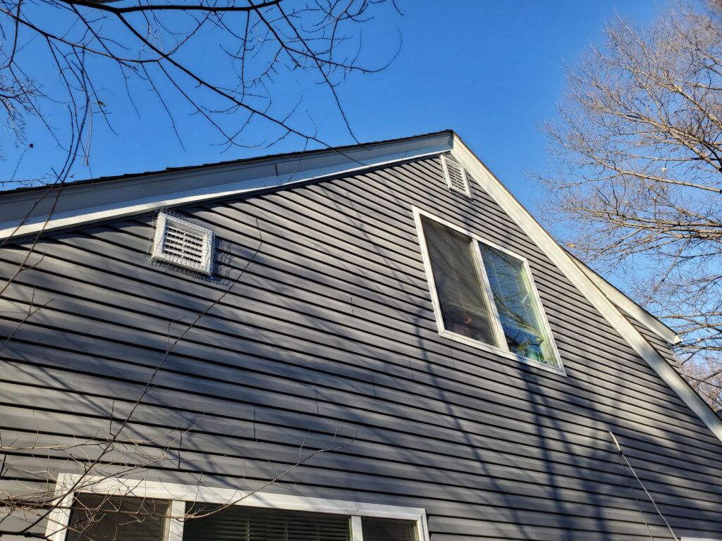 Two white square exterior wall vents on a gray-sided house, both secured with heavy-duty galvanized steel mesh covers.