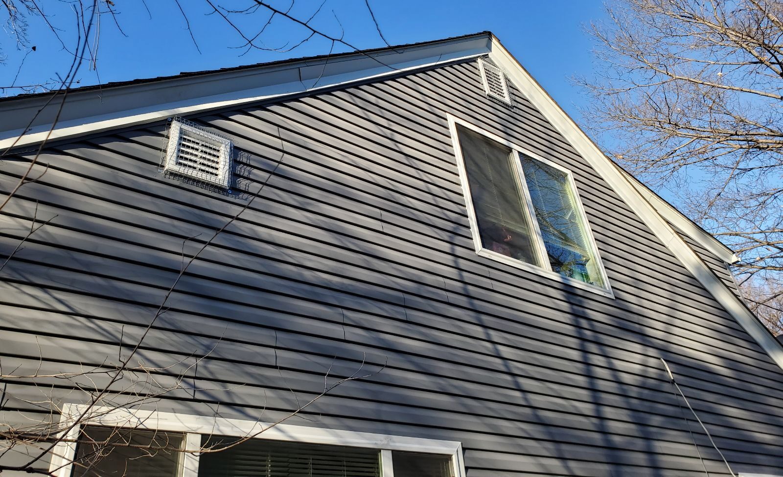 Two white square exterior wall vents on a gray-sided house, both secured with heavy-duty galvanized steel mesh covers.