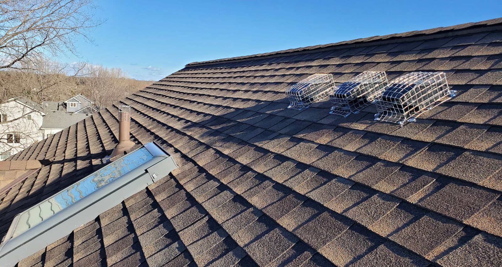 Three roof vents secured with heavy-duty metal mesh wildlife screens on a brown shingled roof under a clear blue sky.