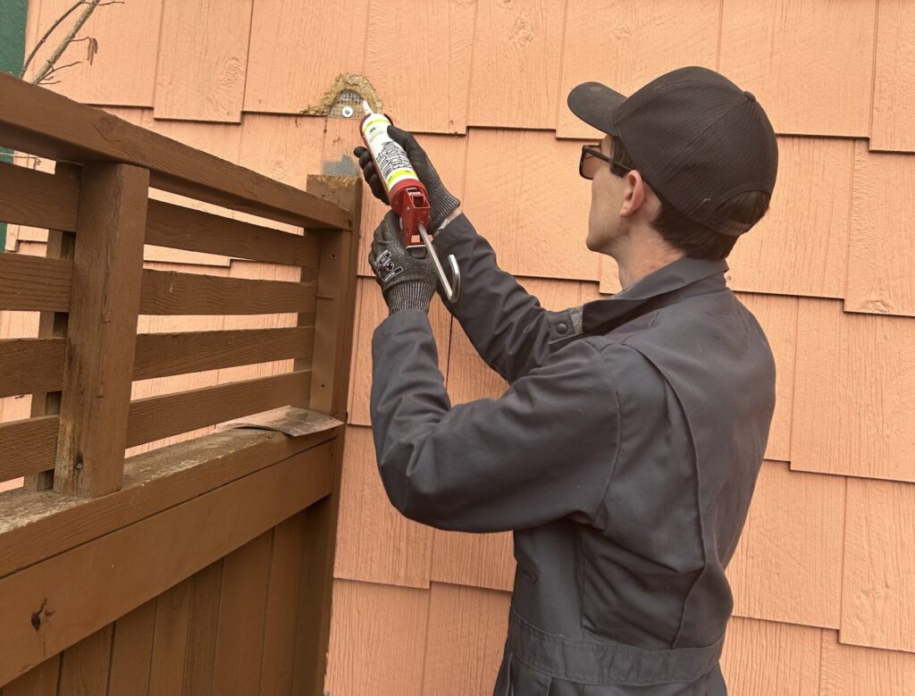 A Skedaddle technician in a grey jumpsuit and work gloves using a caulking gun to seal a small entry hole in orange cedar shingle siding with specialized wildlife sealant.