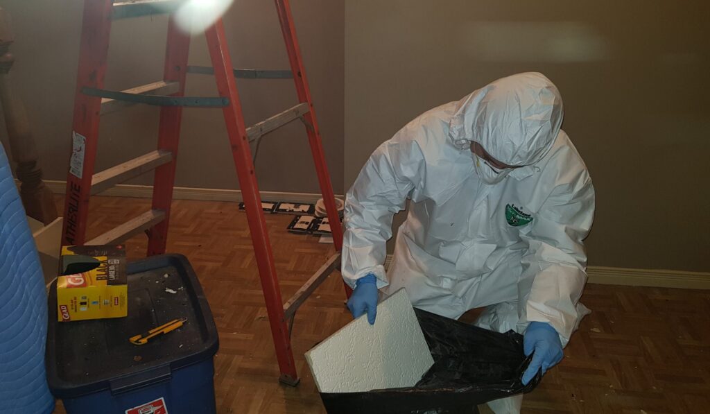A Skedaddle technician in a full white hazmat suit, respirator, and blue gloves disposes of contaminated ceiling tiles into a heavy-duty black waste bag.