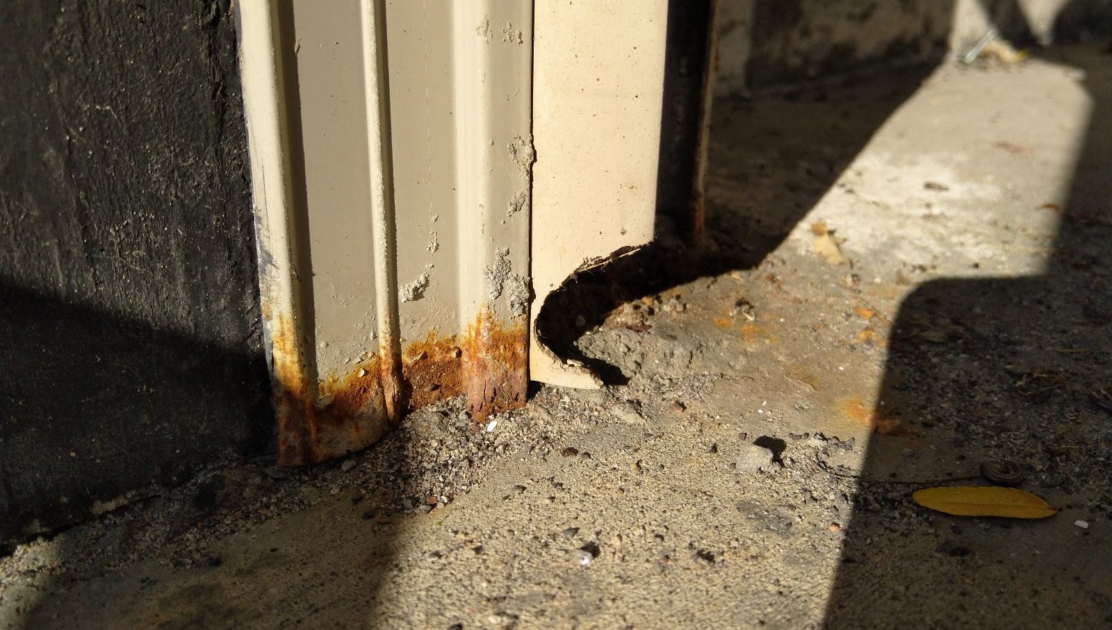 A close-up of a rusted beige metal door frame showing a small, chewed-out hole at the base where it meets the concrete floor, indicating a wildlife entry point.