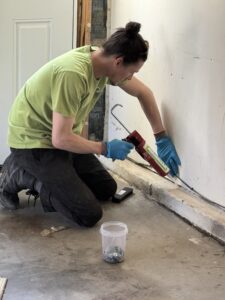 A Skedaddle Wildlife Control technician in a green branded t-shirt and blue gloves uses a caulking gun to seal a gap along a garage wall and floor to prevent animal entry.