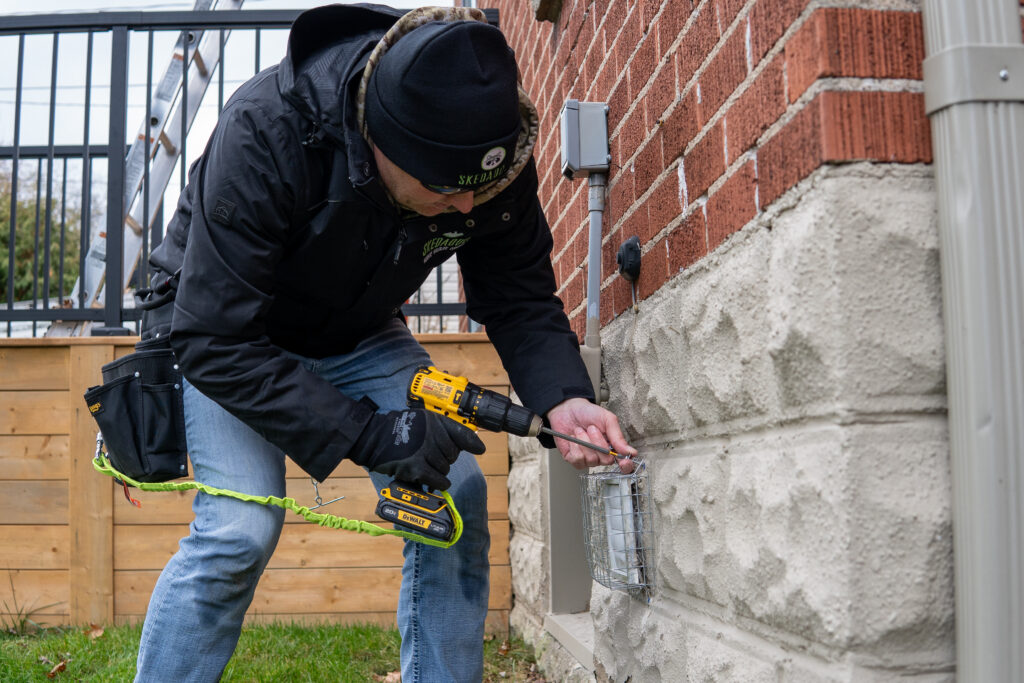A Skedaddle technician in professional gear using a power drill to secure a heavy-duty wire mesh bird guard over an exterior wall vent.