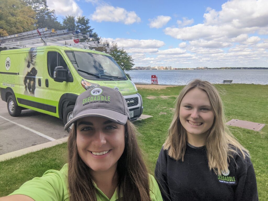 Two smiling female Skedaddle Humane Wildlife Control technicians posing for a selfie in front of a branded service van near a lake.
