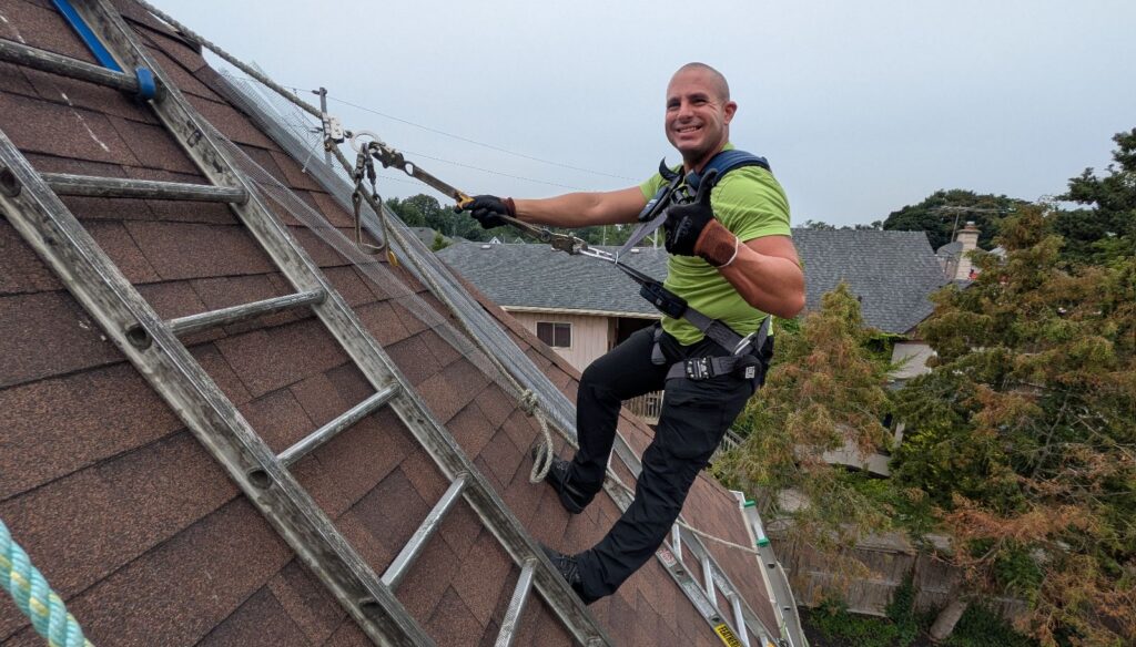 A smiling Skedaddle Pittsburgh technician in a green shirt and safety harness giving a thumbs up while working on a steep roof.