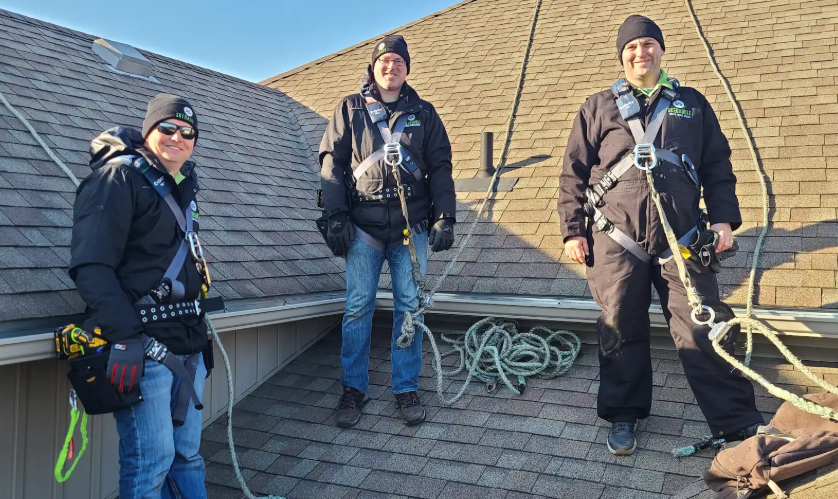 Three Skedaddle Humane Wildlife Control technicians wearing professional uniforms and full safety harnesses while standing on a residential shingle roof.