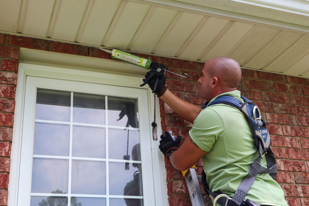 A Skedaddle technician wearing a safety harness uses a caulking gun to seal gaps between a brick wall and white soffit to prevent wildlife entry.