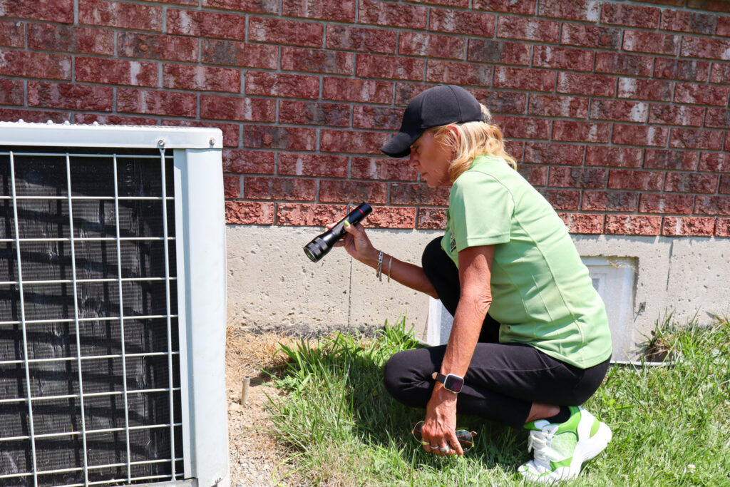 A Skedaddle technician in a green uniform uses a flashlight to inspect the foundation and exterior AC unit of a brick house for signs of wildlife entry.