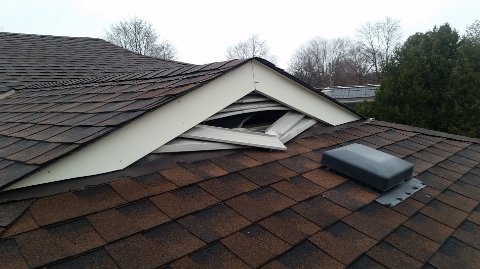 A close-up of a brown shingled roof where the white aluminum soffit under a dormer peak has been torn open and mangled by a raccoon to gain attic access.