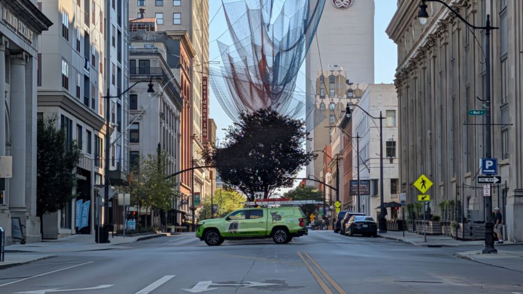 A bright green Skedaddle Humane Wildlife Control service truck driving through a busy metropolitan downtown street with tall office buildings and a large decorative net installation above.