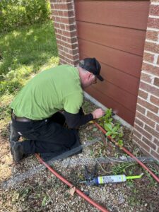 wildlife exclusion sealant foundation repair A Skedaddle technician in a lime green shirt kneeling to apply specialized wildlife exclusion sealant and mesh along a home's foundation.
