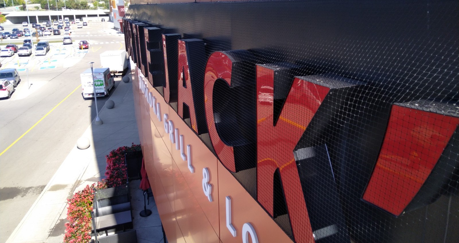 A high-angle, side-profile view of a large "Kelsey's" restaurant sign covered in fine black bird netting to prevent birds from nesting in the channel letters.