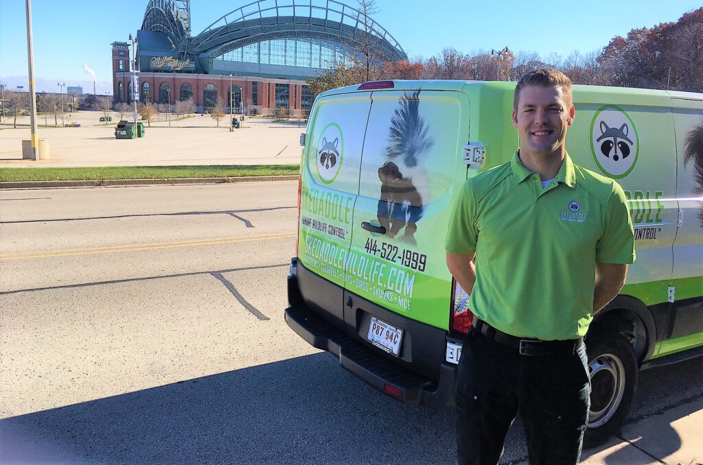 Marcus Mueller stands next to a Skedaddle Humane Wildlife Control van branded with raccoon and skunk graphics, with a large domed stadium in the background.