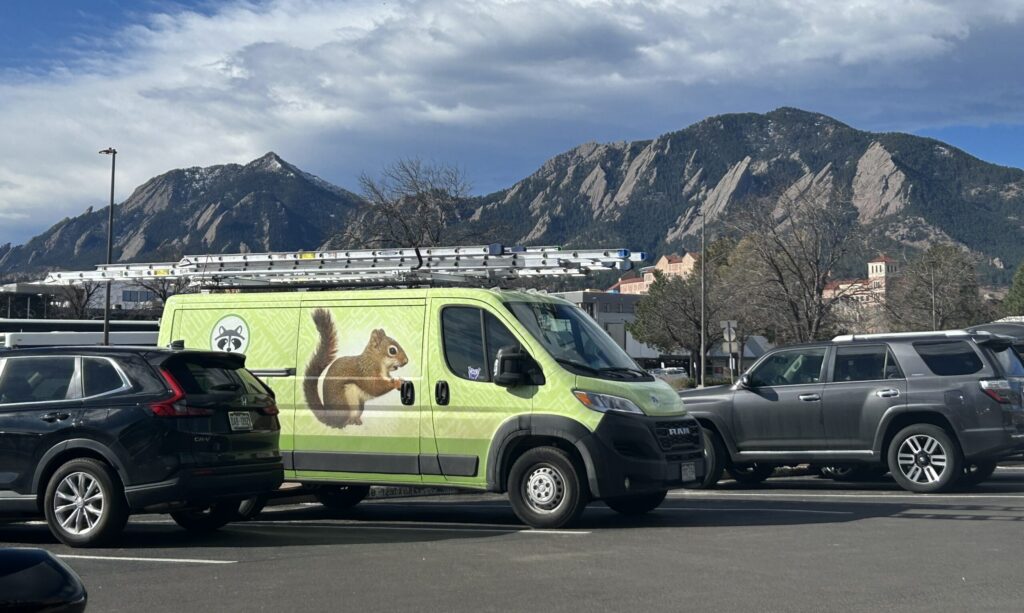 A lime-green Skedaddle RAM service van with a large squirrel graphic parked in a lot, featuring the iconic Flatirons rock formations and a blue sky in the background.