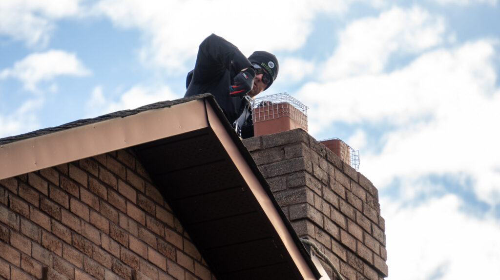 A Skedaddle wildlife technician on a roof installing a heavy-duty wire mesh screen over a brick chimney to prevent animal entry.