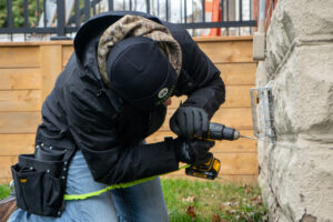 A Skedaddle wildlife technician using a power drill to install a heavy-duty mesh exclusion screen over an exterior wall vent.