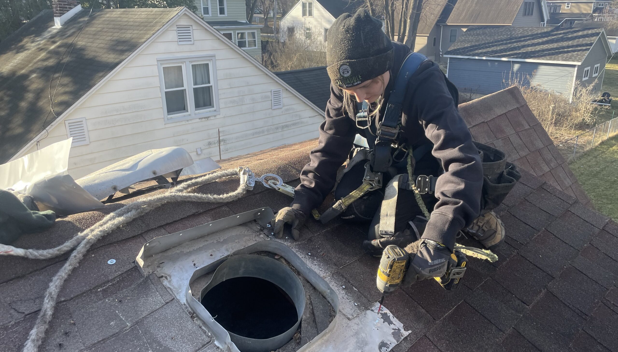 A Skedaddle Humane Wildlife Control technician wearing a safety harness and beanie, using a power drill to secure a protective metal cover over a roof vent to prevent animal entry.