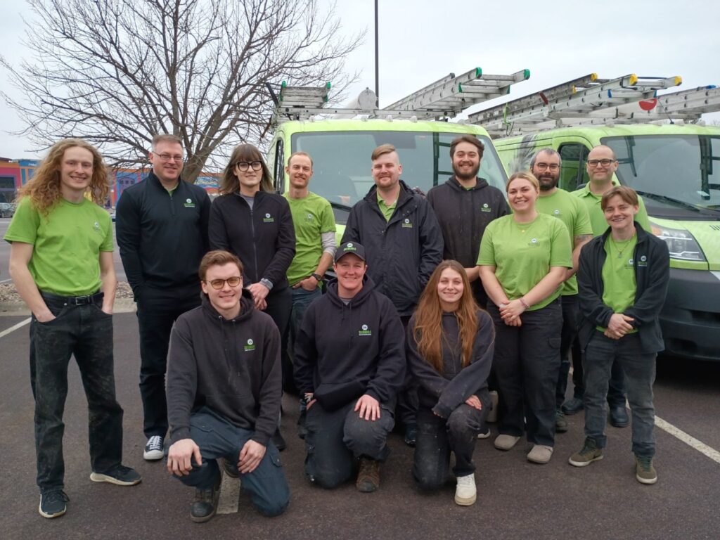 A large group of Skedaddle technicians in branded green and black gear standing in front of two service vans.