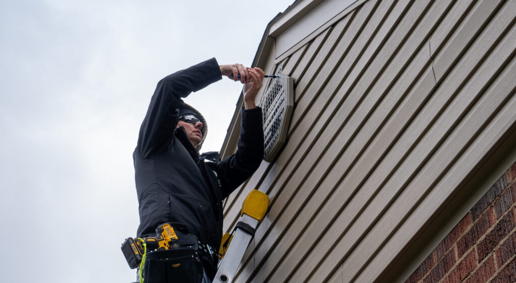 A professional wildlife technician on a ladder installing heavy-duty steel mesh over a house gable vent to prevent animal entry.