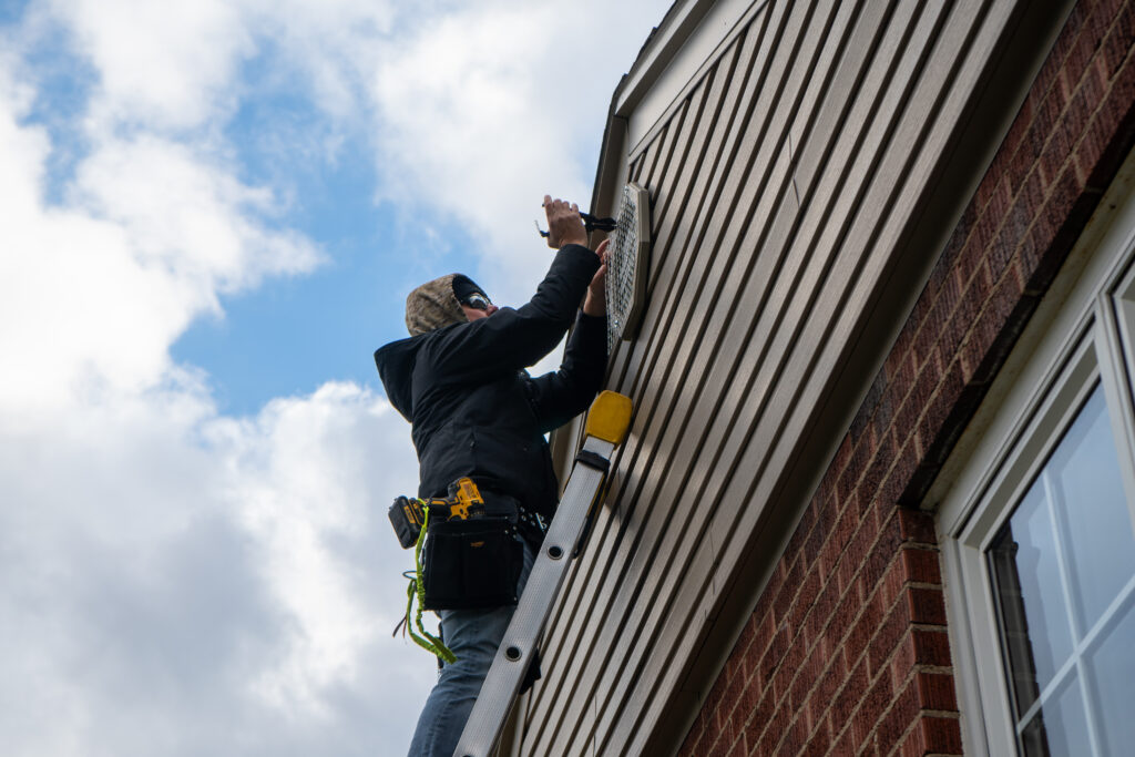 A Skedaddle technician on a ladder installing a heavy-duty mesh screen over a wall vent on a house with tan siding.