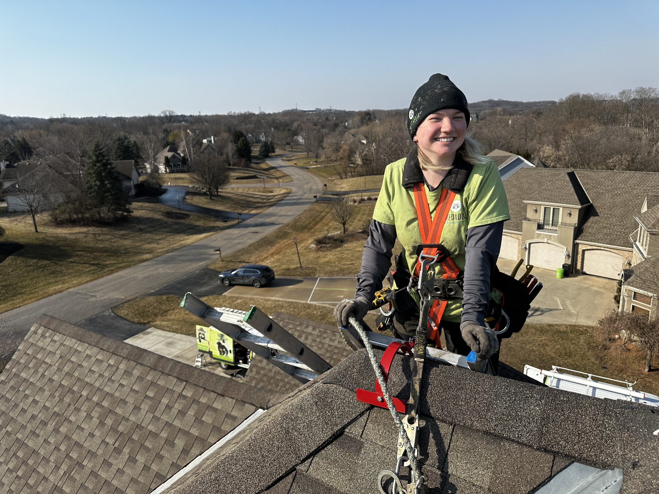 A smiling female wildlife technician in a lime green shirt and safety harness sitting on a shingled rooftop with a suburban neighborhood in the background.