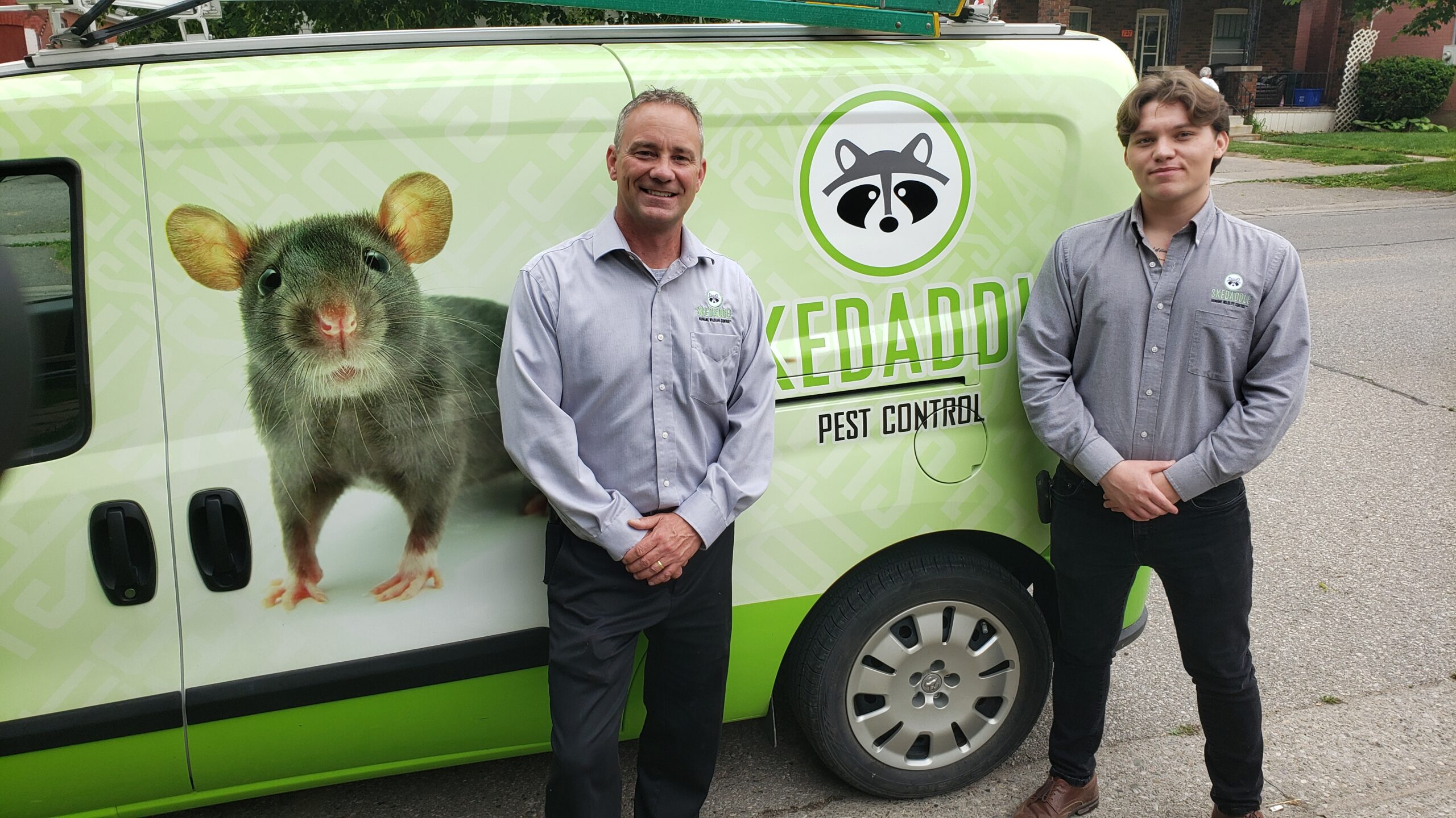 Two Skedaddle Pest Control team members standing proudly next to a green service van featuring a large rat graphic and company branding.