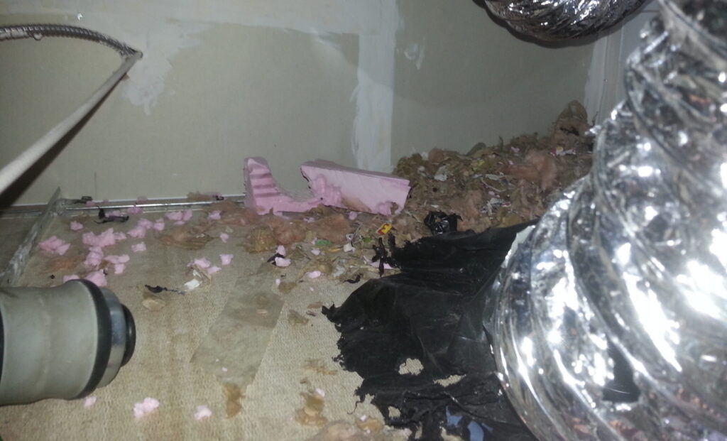 Shredded pink insulation, torn black plastic, and nesting debris scattered in a confined attic space near silver flexible ducting and copper pipes.