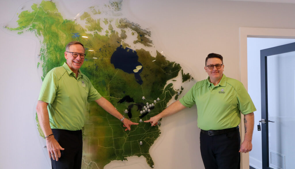 Bill and Geoff, wearing lime green Skedaddle shirts, stand in an office and point to various franchise locations on a large wall-mounted map of North America.
