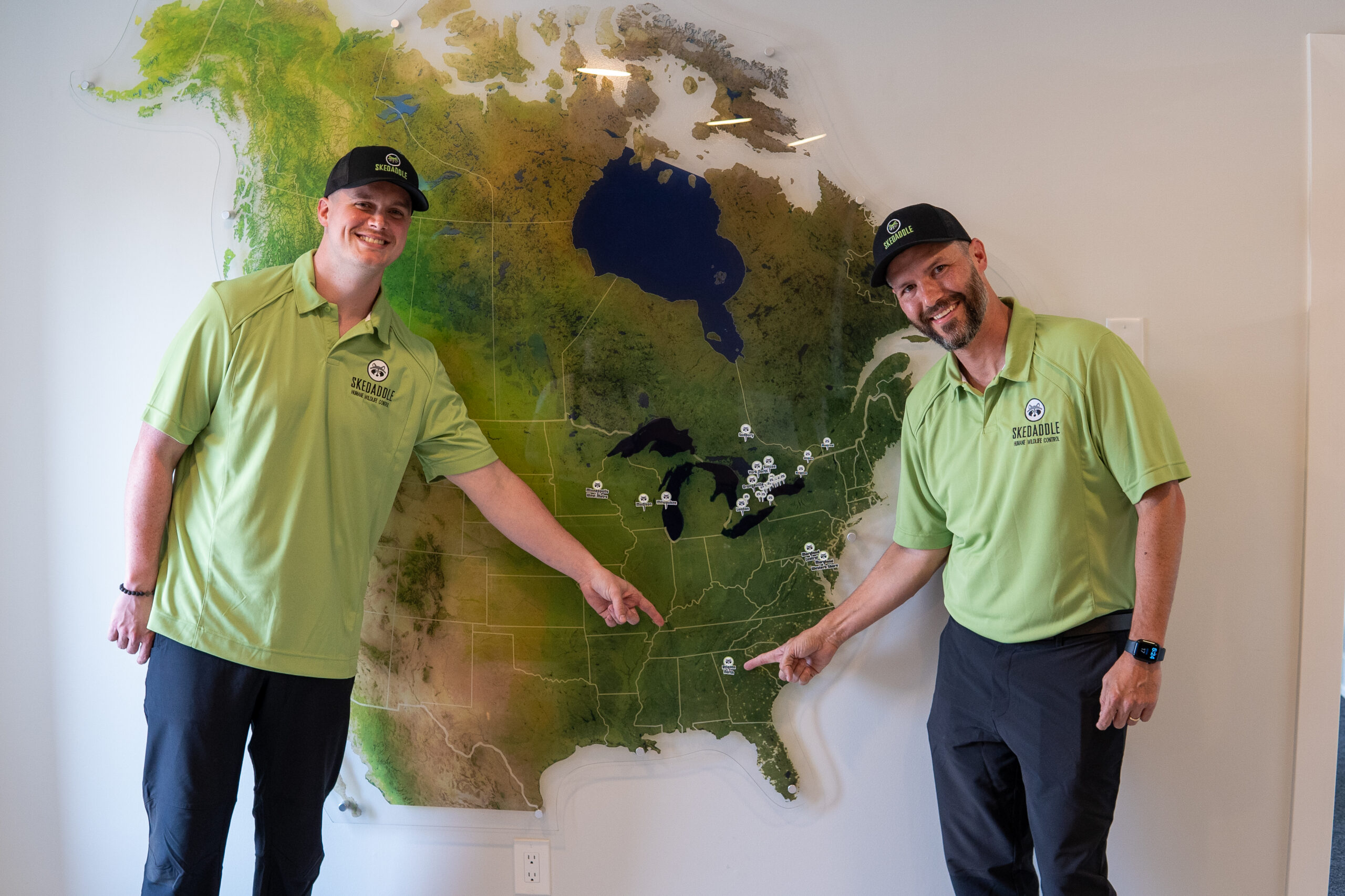 Two smiling Skedaddle Wildlife Control team members in green polo shirts pointing at franchise locations on a large North American map wall display.