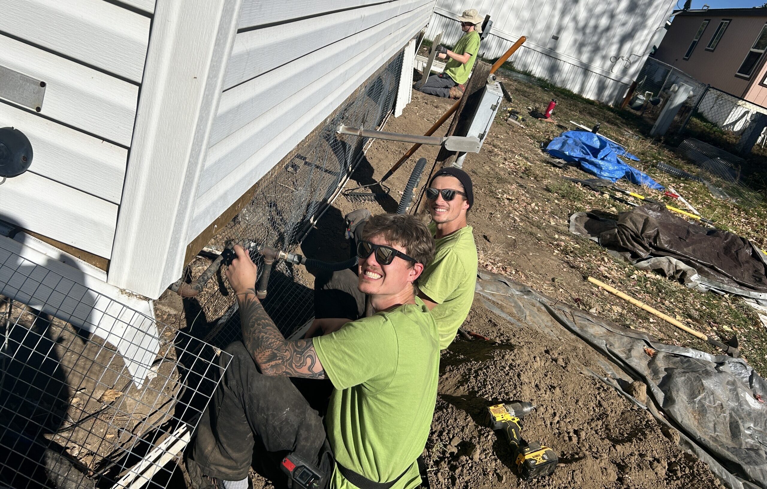 Two Skedaddle technicians in green shirts smiling while installing a heavy-duty wire mesh screen to the base of a home in Denver to prevent wildlife entry.