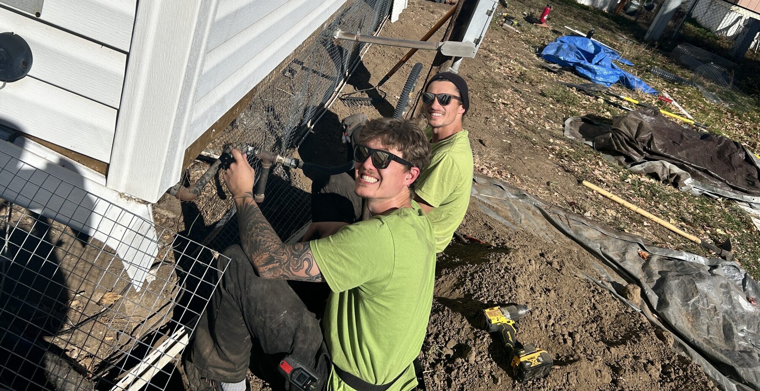 Two Skedaddle technicians in green shirts smiling while installing a heavy-duty wire mesh screen to the base of a home in Denver to prevent wildlife entry.