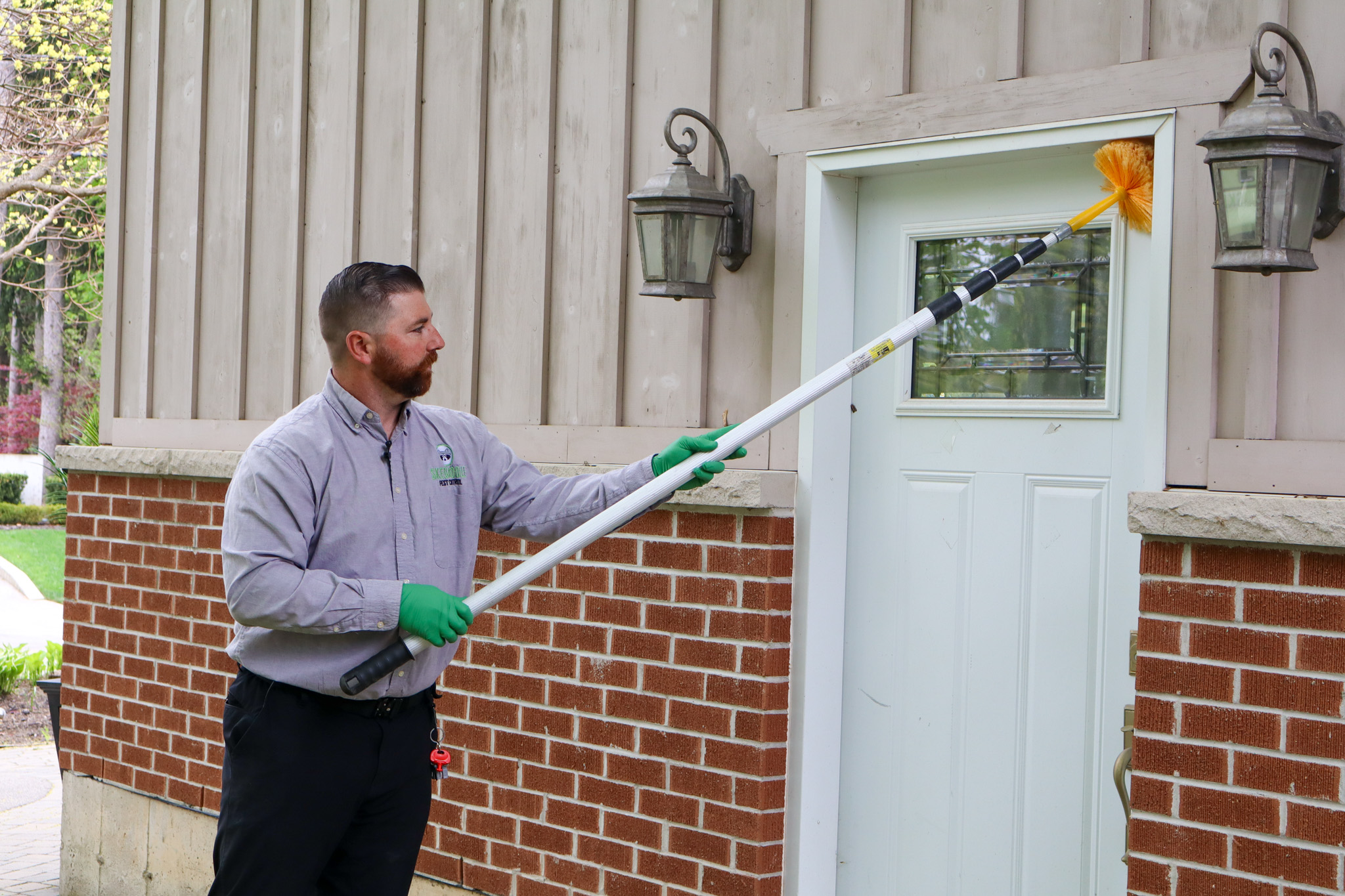 A Skedaddle Humane Wildlife Control technician wearing green safety gloves using a long telescopic brush pole to sweep the top corner of a residential front door frame.